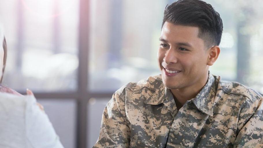 A person in military uniform sitting opposite another individual, indoors with a bright window background.